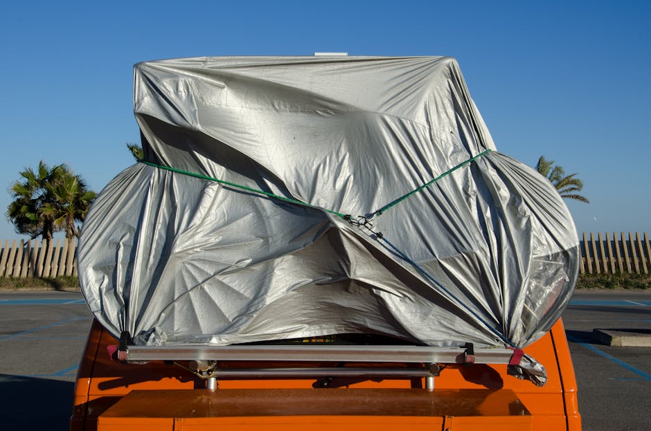 An orange vehicle parked outdoors covered in a protective tarp under a clear blue sky.